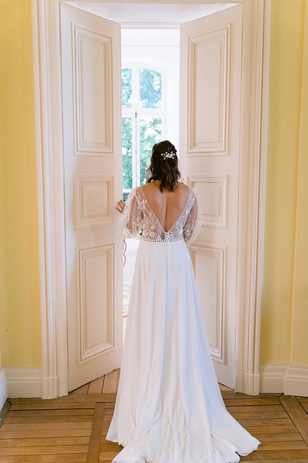 Vue de dos, la mariée se tient devant une fenêtre ouverte, sa robe blanche à dos en V déployée sur le parquet.