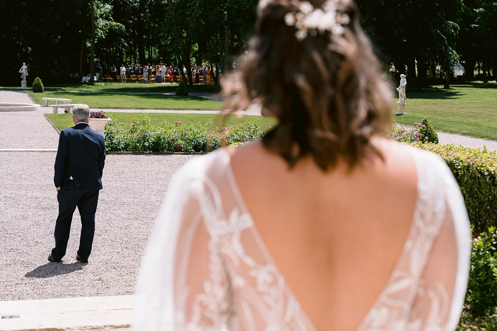 La mariée est vue de dos au premier plan tandis qu’un homme en costume l’attend plus loin dans la cour.