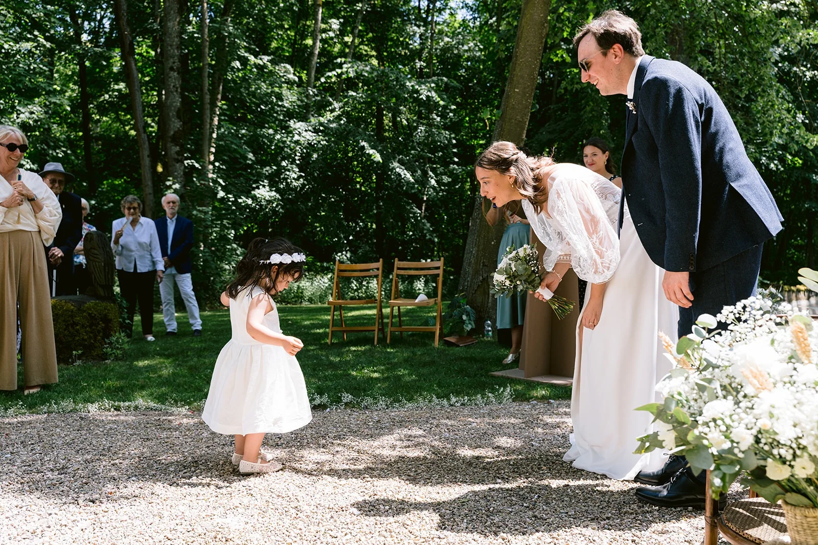 Les mariés se penchent vers un enfant en robe blanche qui s’approche d’eux pendant la cérémonie sous les arbres.