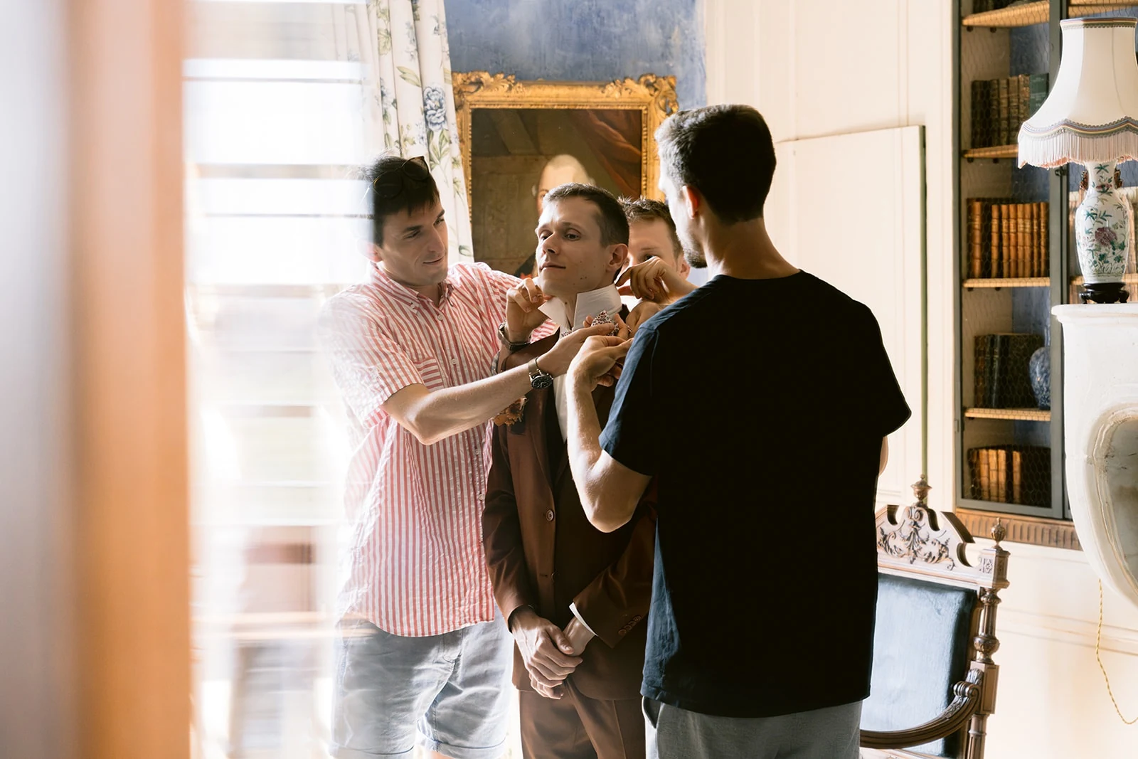 Plusieurs personnes ajustent le col et le nœud papillon du marié, vêtu de son costume brun.