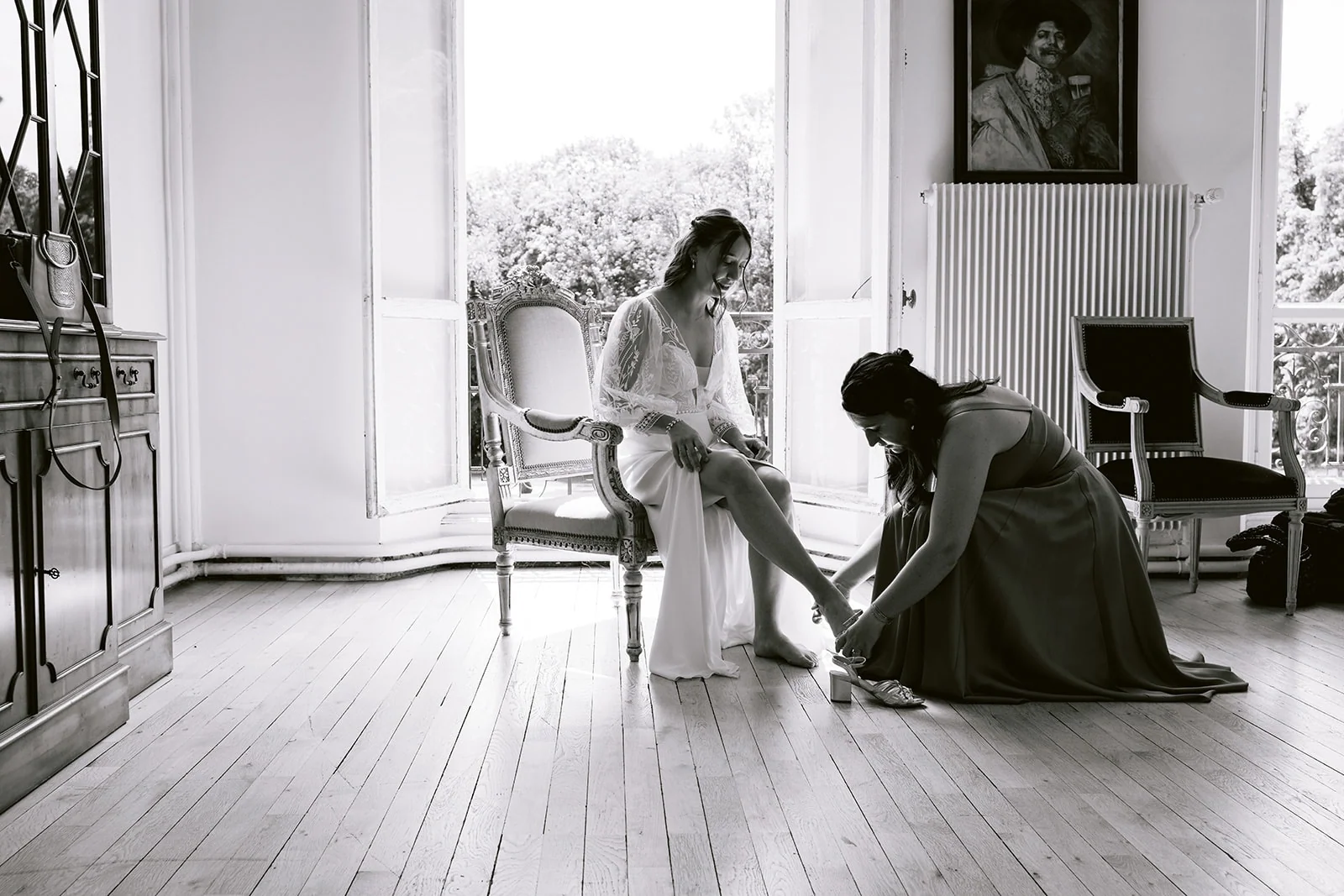En noir et blanc, une femme agenouillée ajuste la chaussure de la mariée, assise près d’une fenêtre ouverte.