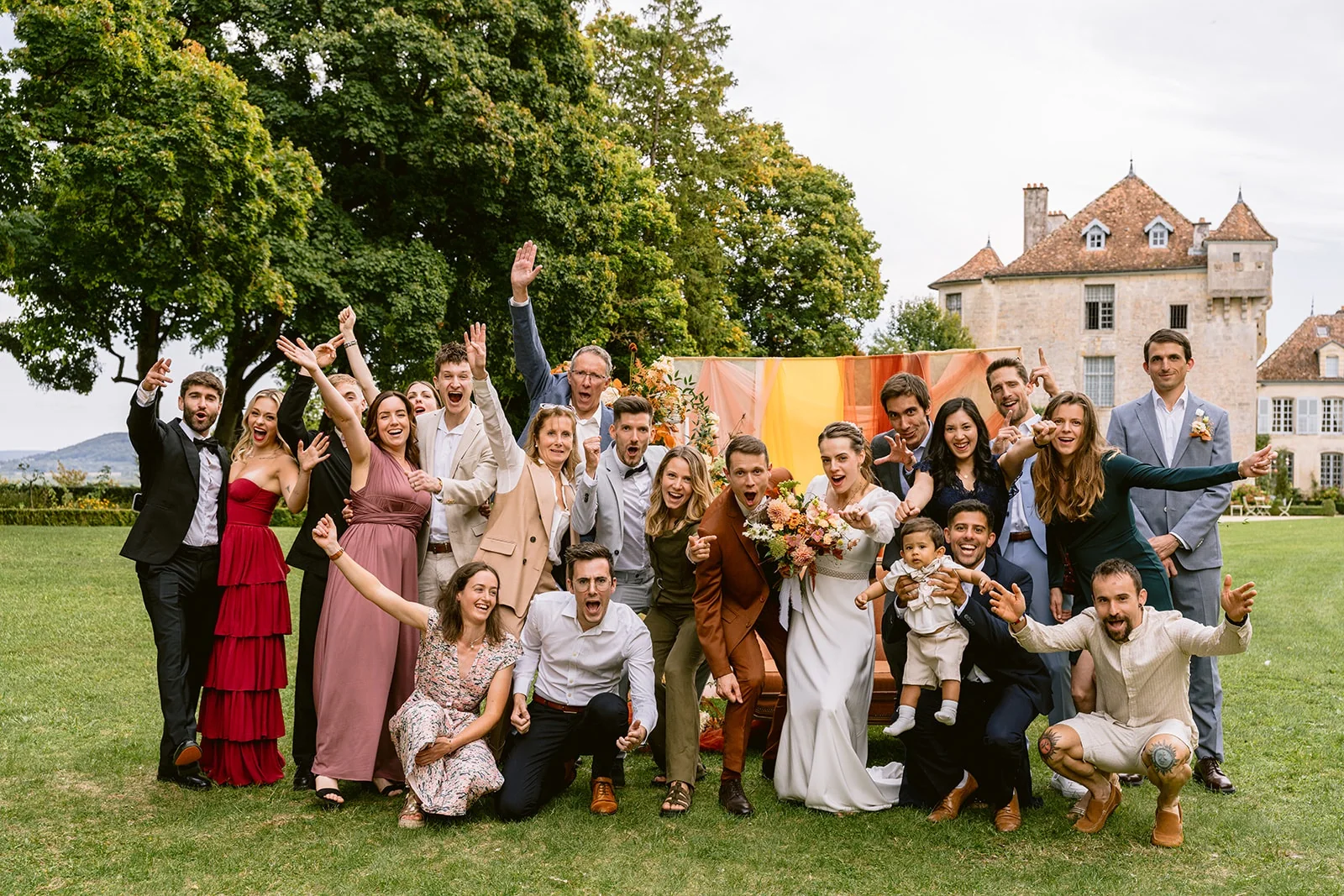 Les mariés posent sur la pelouse avec un groupe d’invités qui lèvent les bras devant une demeure en pierre.