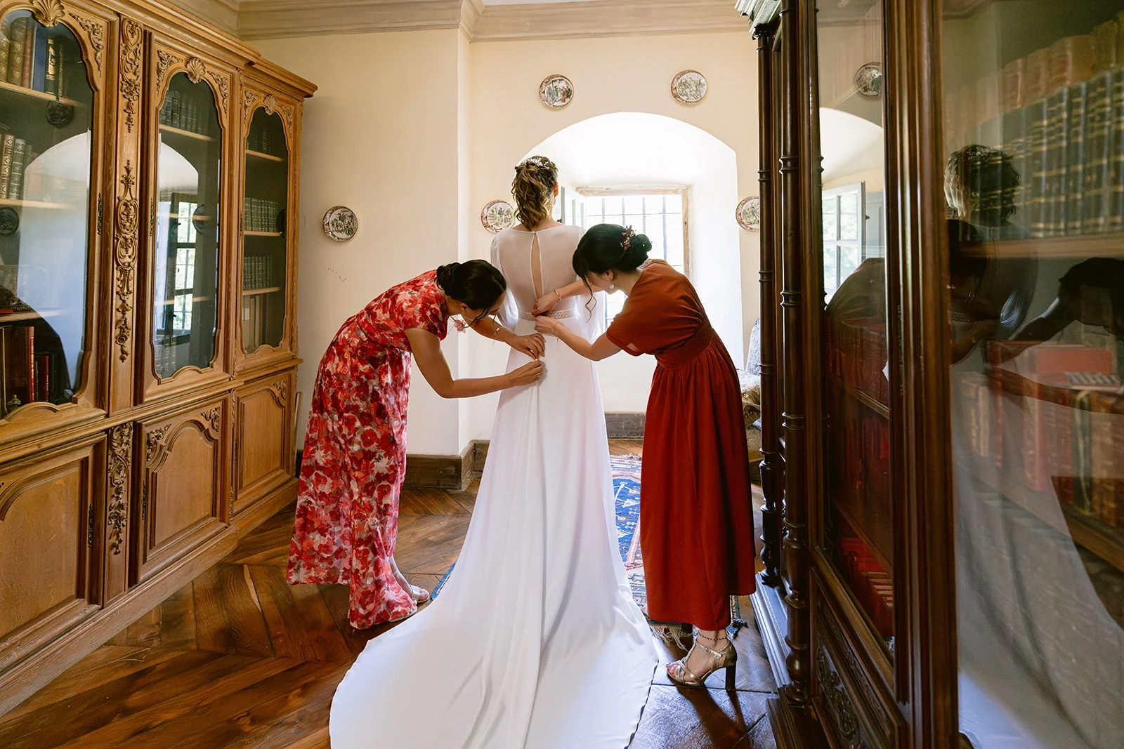 Deux femmes ferment le dos de la robe de mariée, dont la longue traîne s’étale entre deux bibliothèques.
