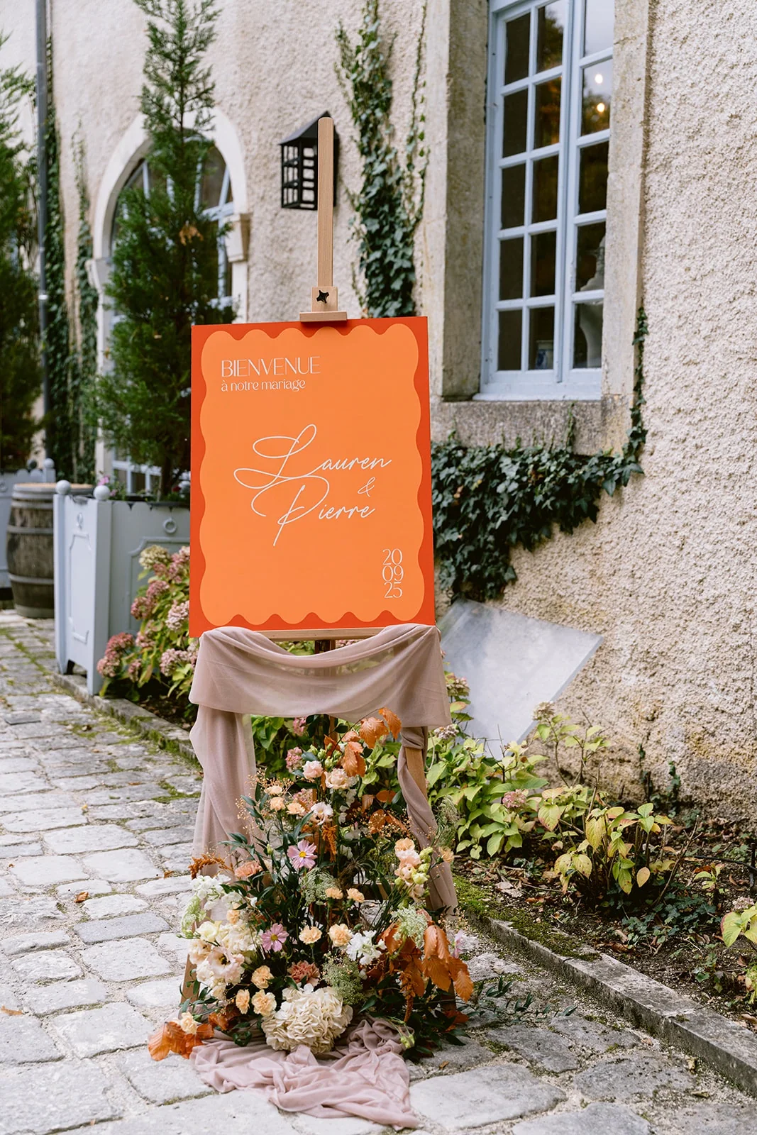 Un panneau d’accueil orangé repose sur un support drapé, avec des fleurs au pied d’une façade ancienne.