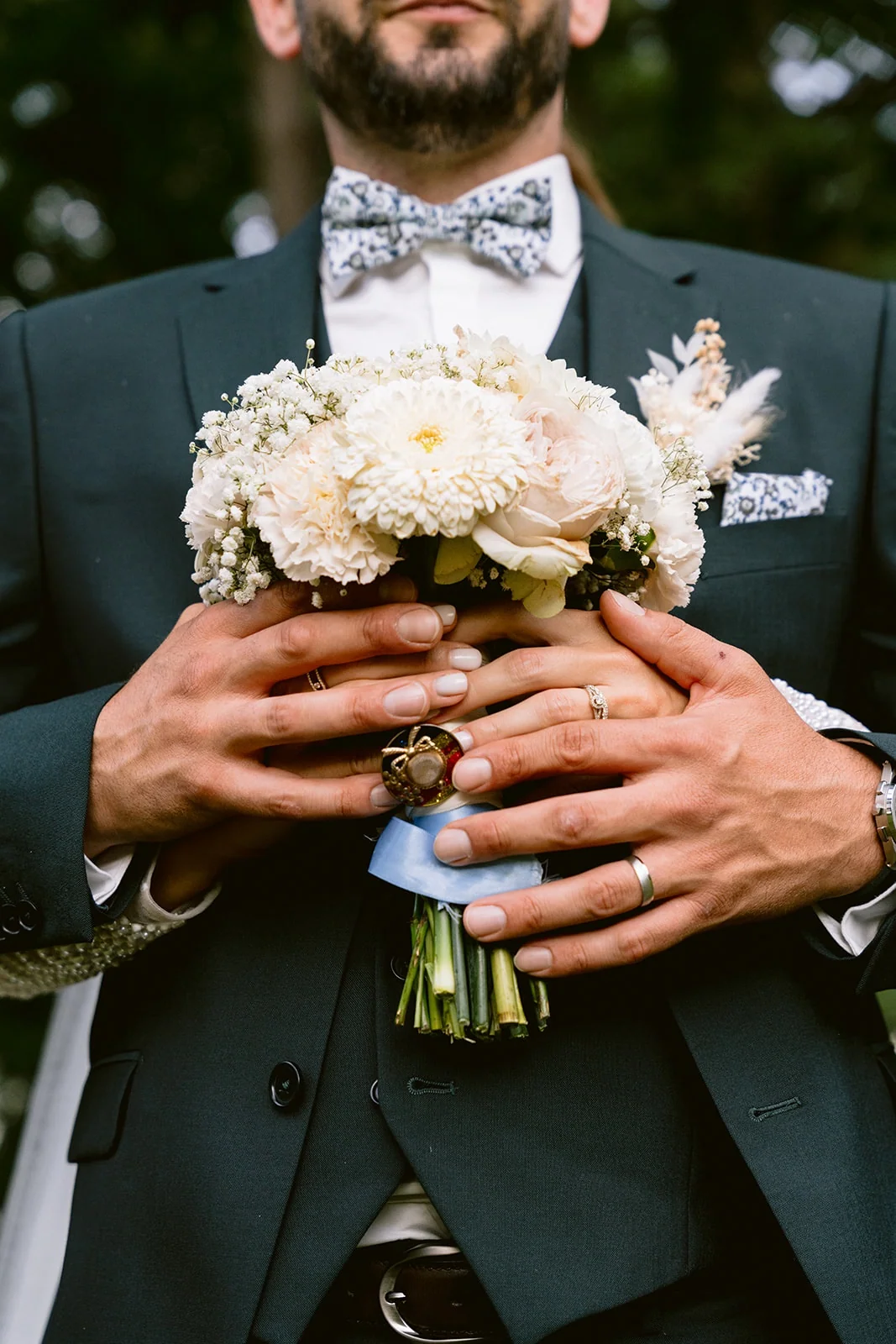 Les mains des mariés se rejoignent autour du bouquet devant le costume sombre.