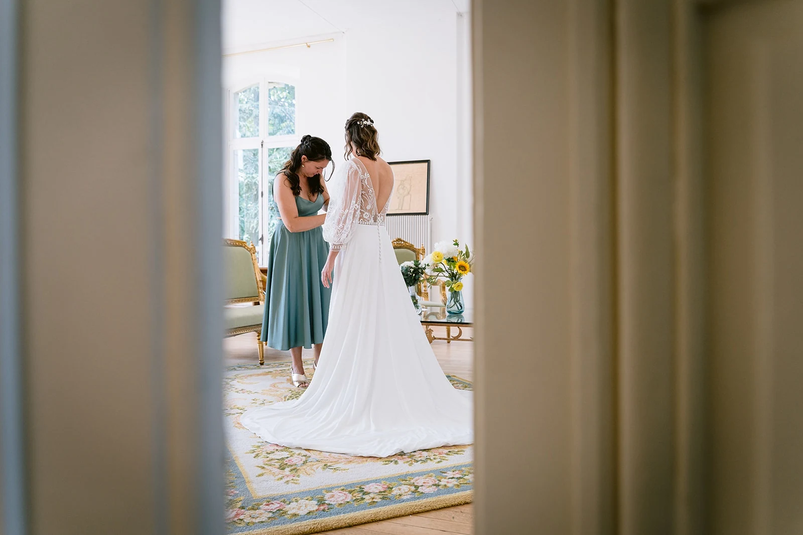 Vues à travers une porte, la mariée et une femme en robe verte ajustent sa tenue près d’une fenêtre.