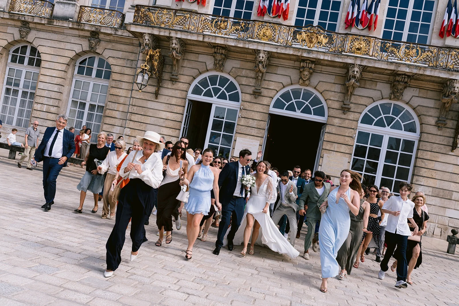 Les mariés et leurs invités traversent une place pavée devant un bâtiment aux grandes fenêtres et aux drapeaux français.