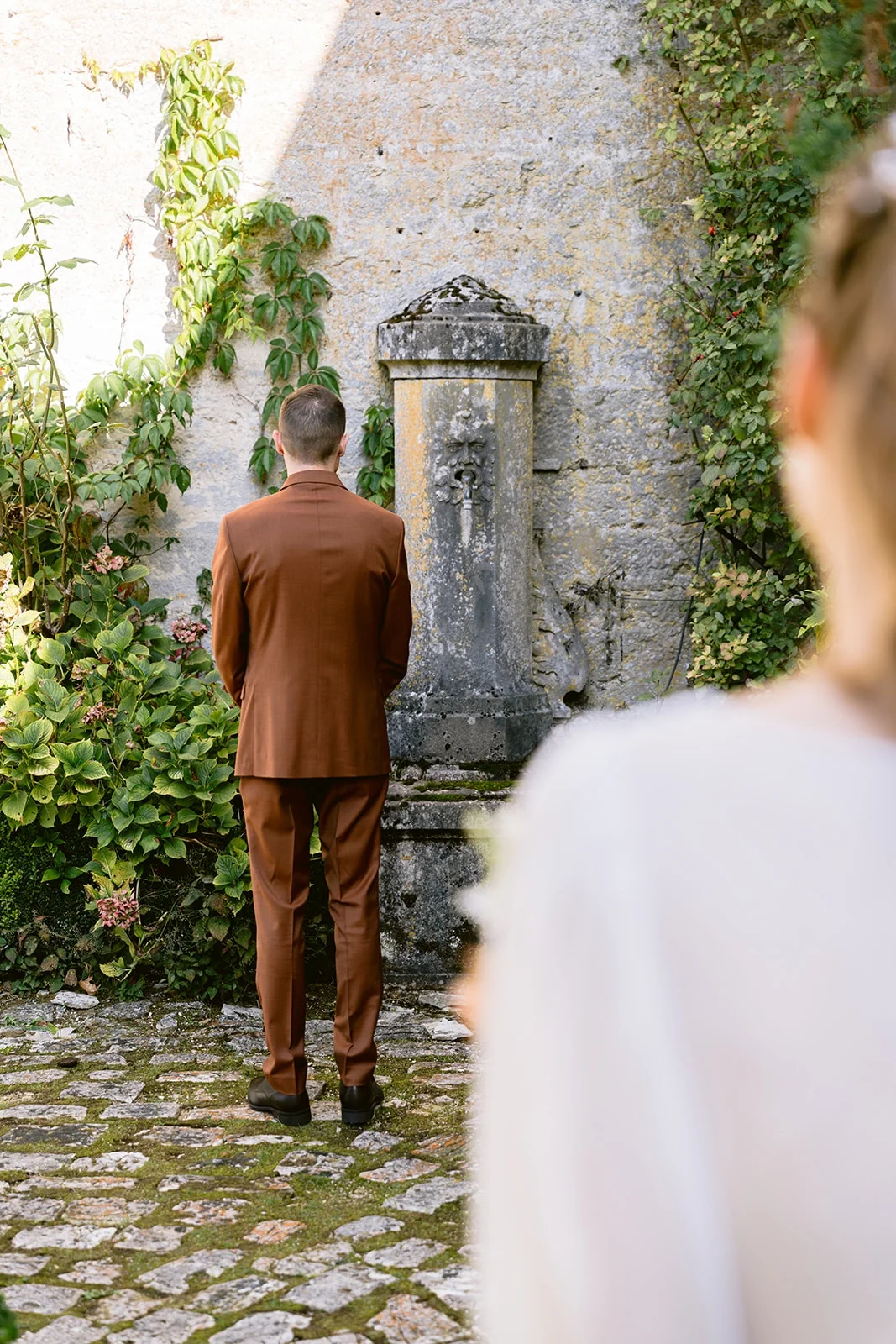 Le marié en costume brun se tient de dos devant une fontaine en pierre, la mariée floue au premier plan.