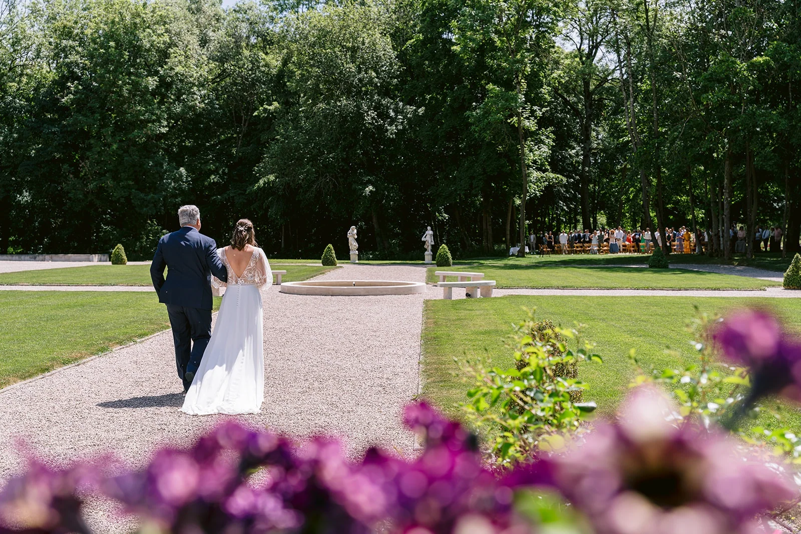 Vus de dos, la mariée et un homme en costume marchent sur une allée de gravier au milieu d’un parc fleuri.