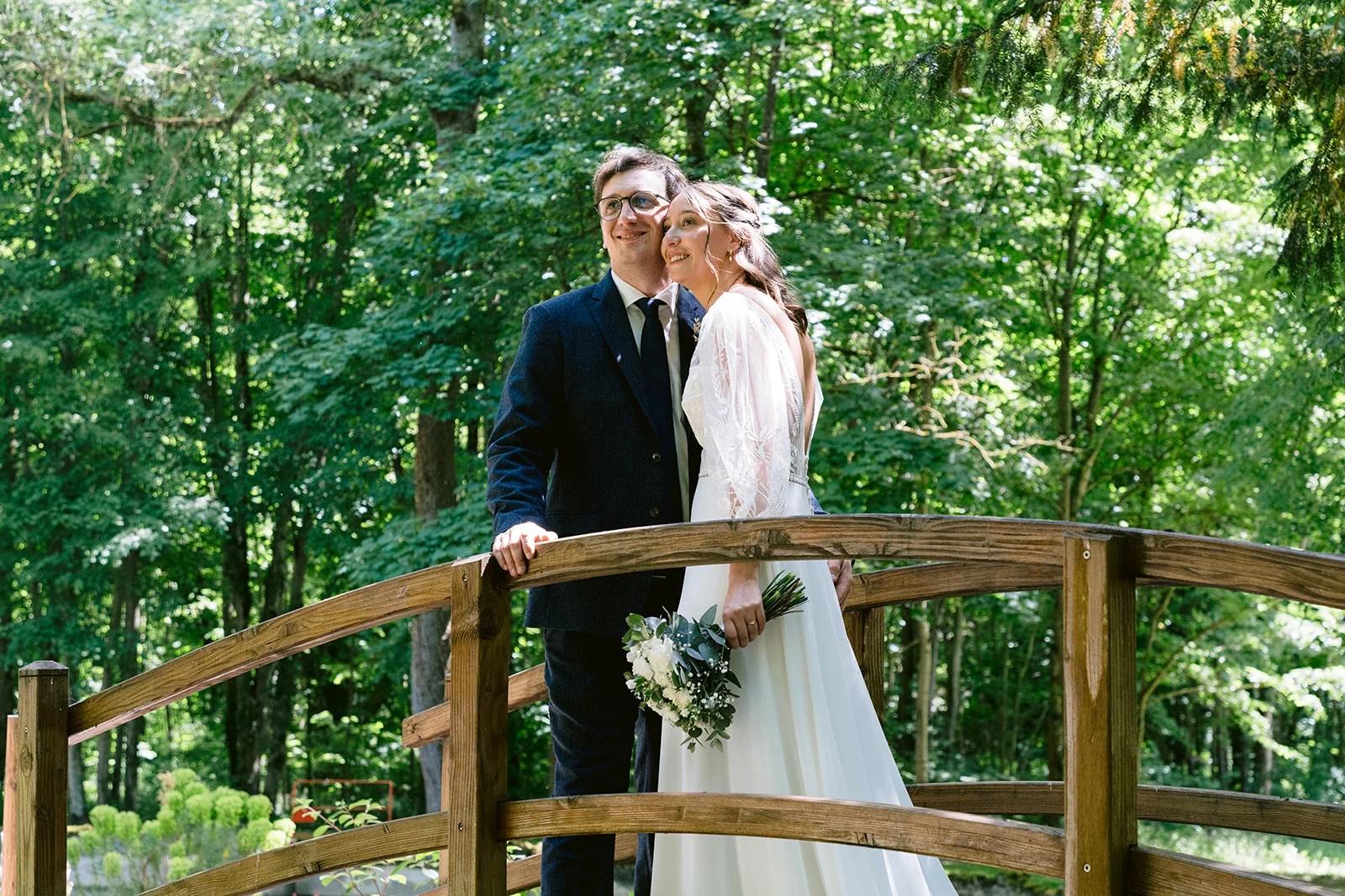 La mariée pose la tête contre celle du marié sur un petit pont de bois entouré d’arbres.