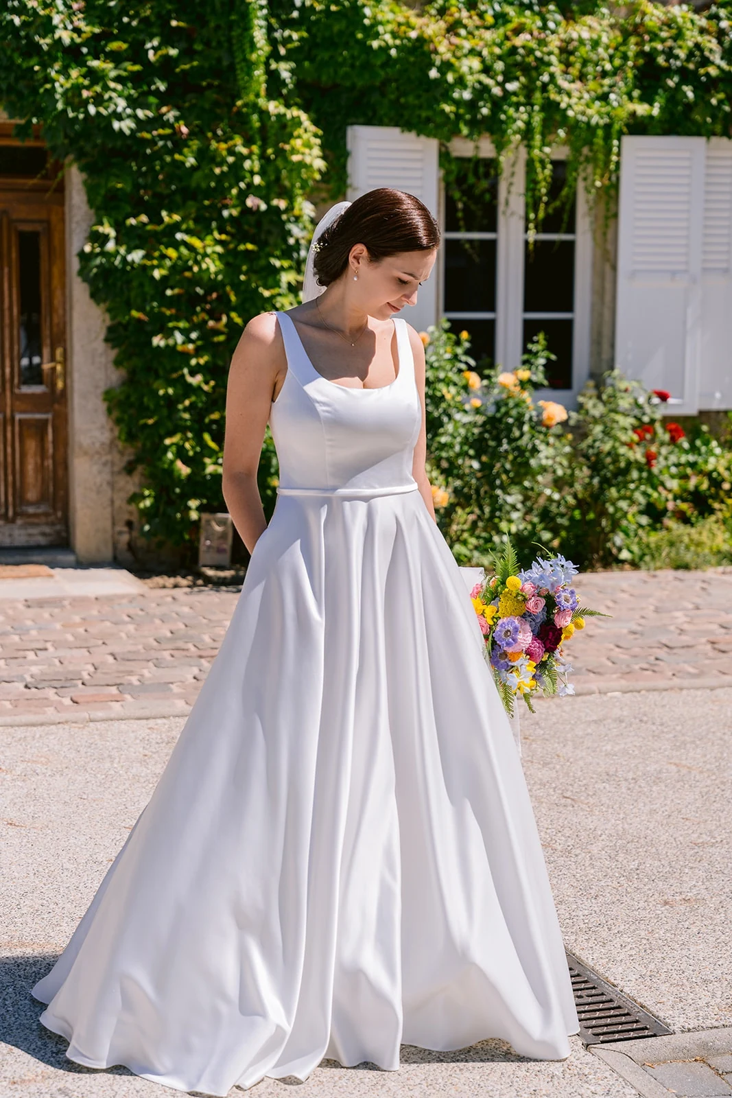 Une mariée tient un bouquet coloré devant une façade couverte de verdure.