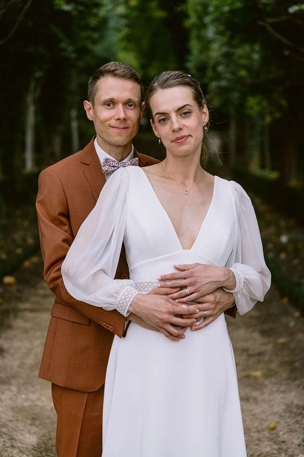 Le marié enlace la mariée par la taille, tous deux face à l’objectif devant des arbres.
