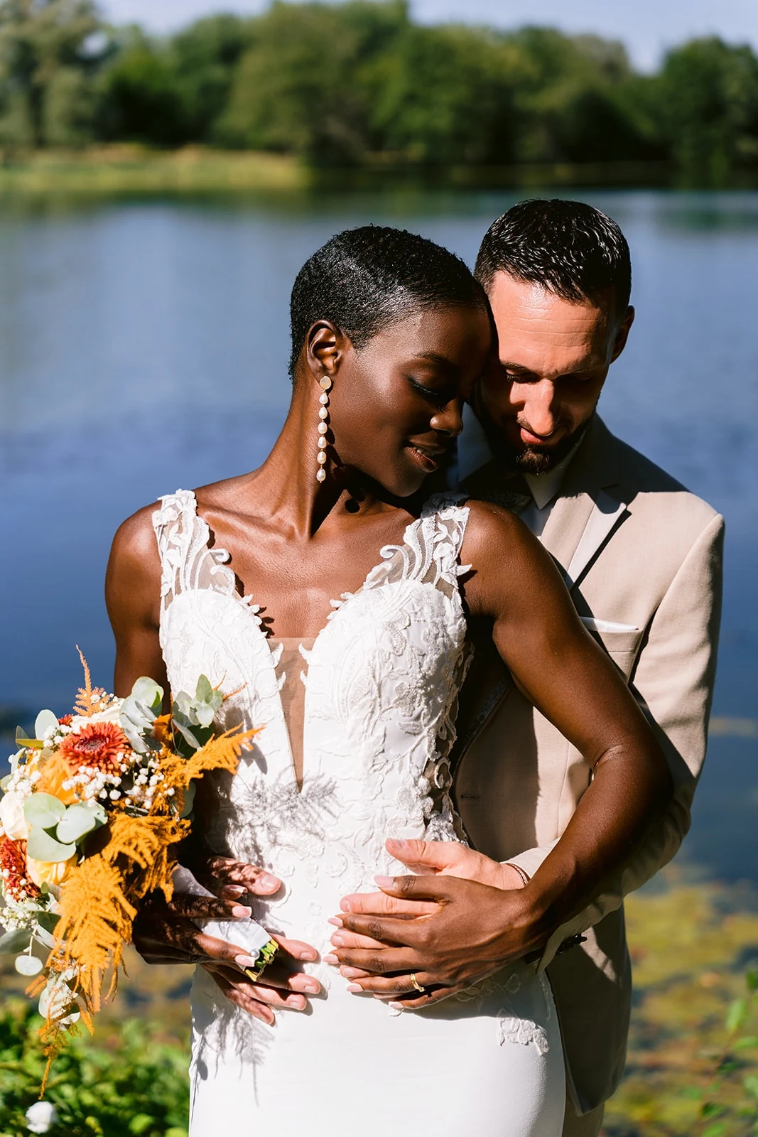 Les mariés enlacés au bord de l’eau, la mariée tenant un bouquet aux fleurs jaunes et blanches.
