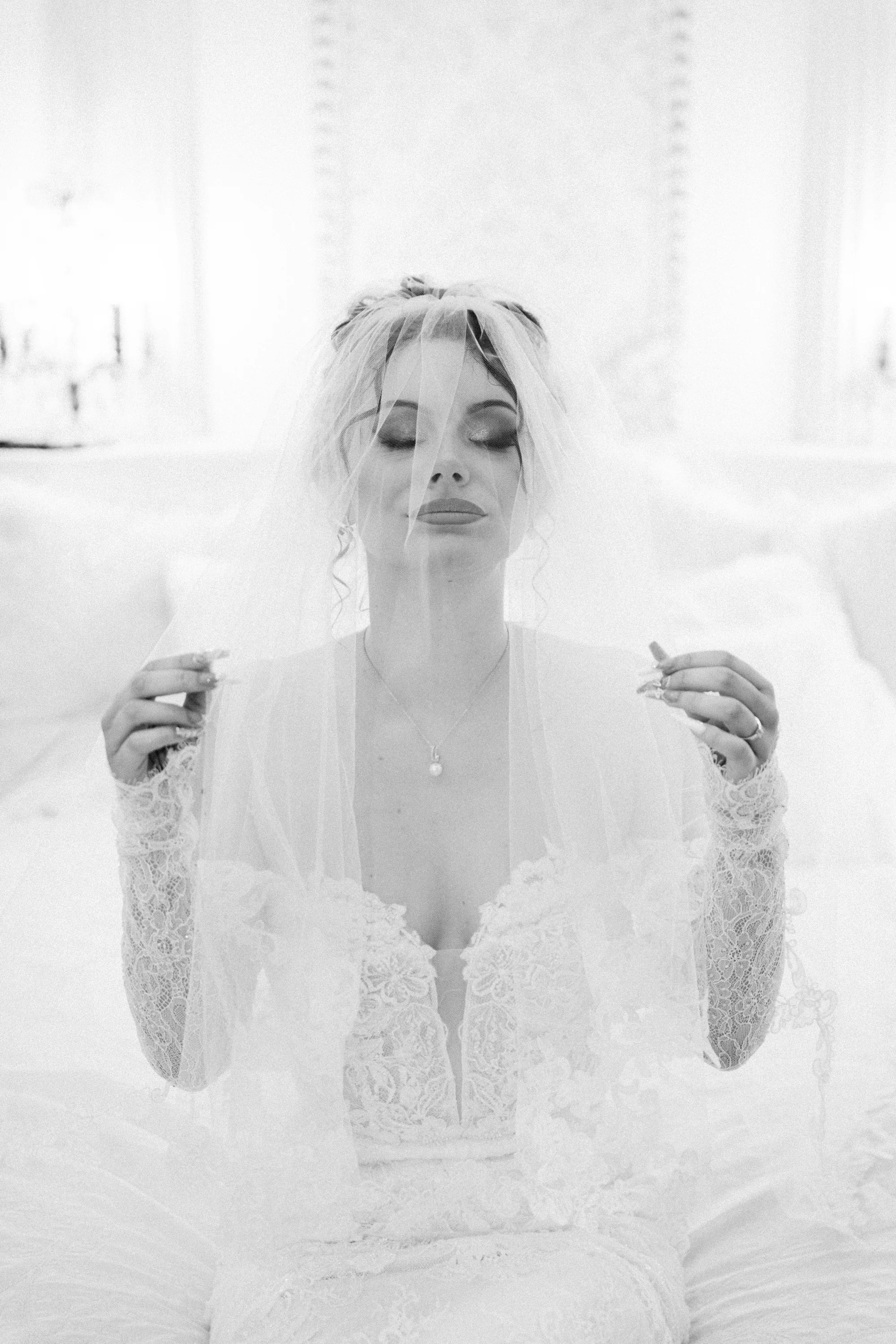 La mariée tient son voile devant son visage, dans une lumière douce en noir et blanc.