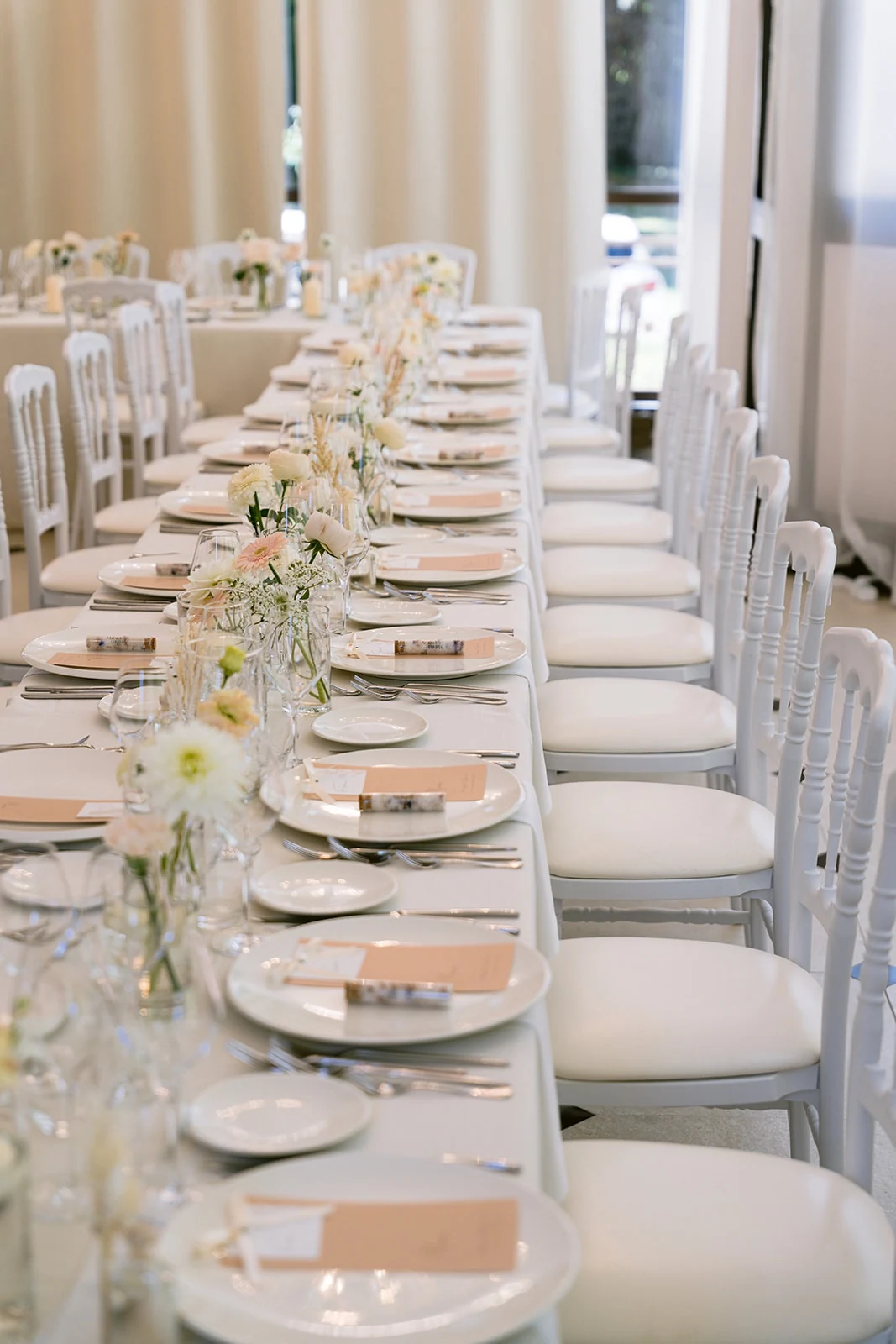 Une longue table blanche est décorée de fleurs et de verres alignés.