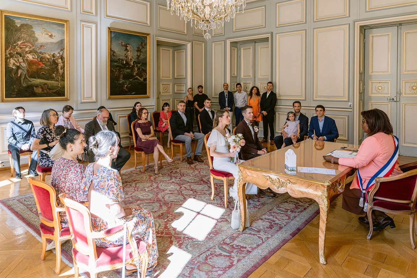 Les mariés et leurs invités assistent à une cérémonie civile dans un salon aux murs ornés de tableaux.