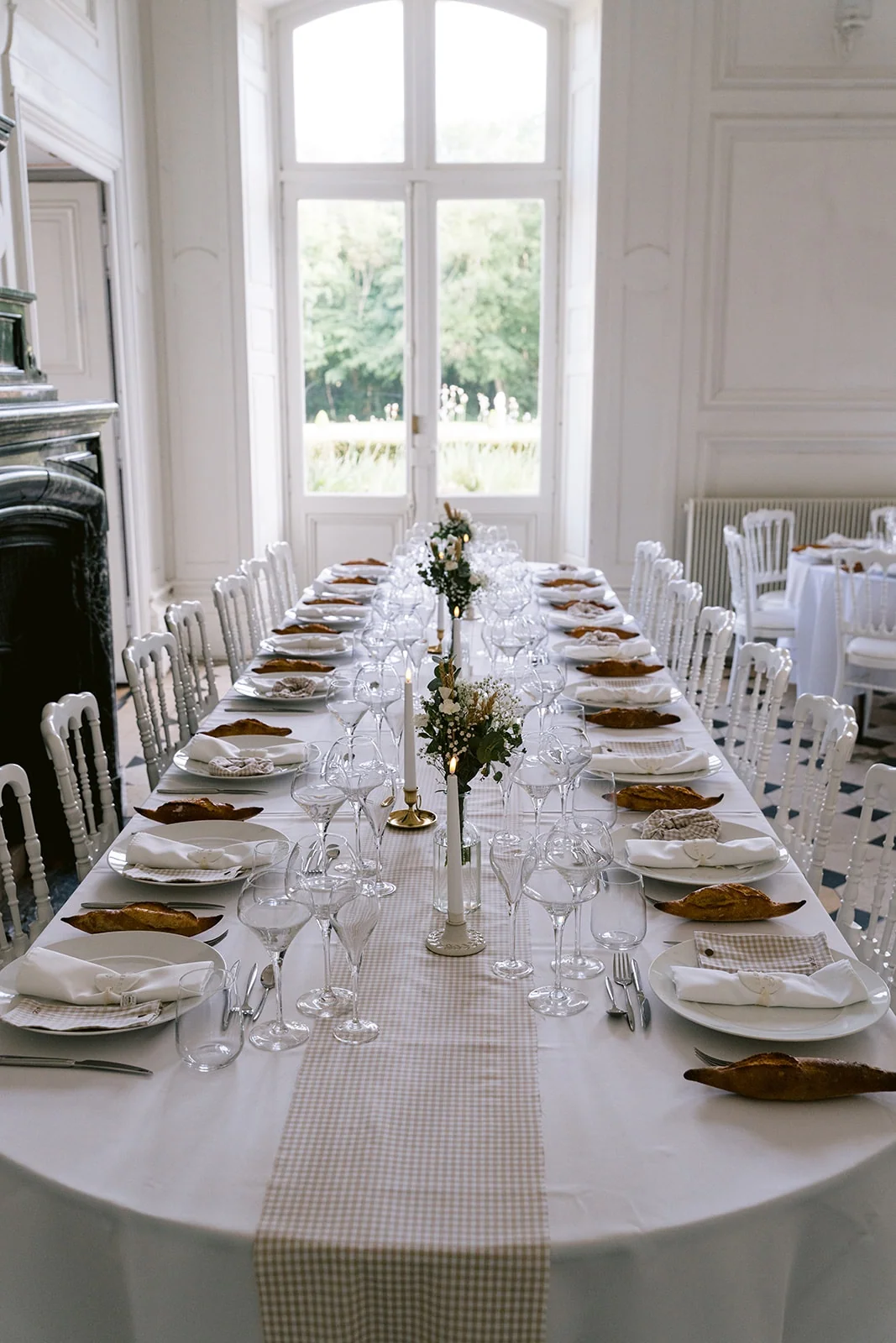 Une longue table de réception, dressée de vaisselle blanche, de verres et de petits bouquets, s’étend devant une grande fenêtre.