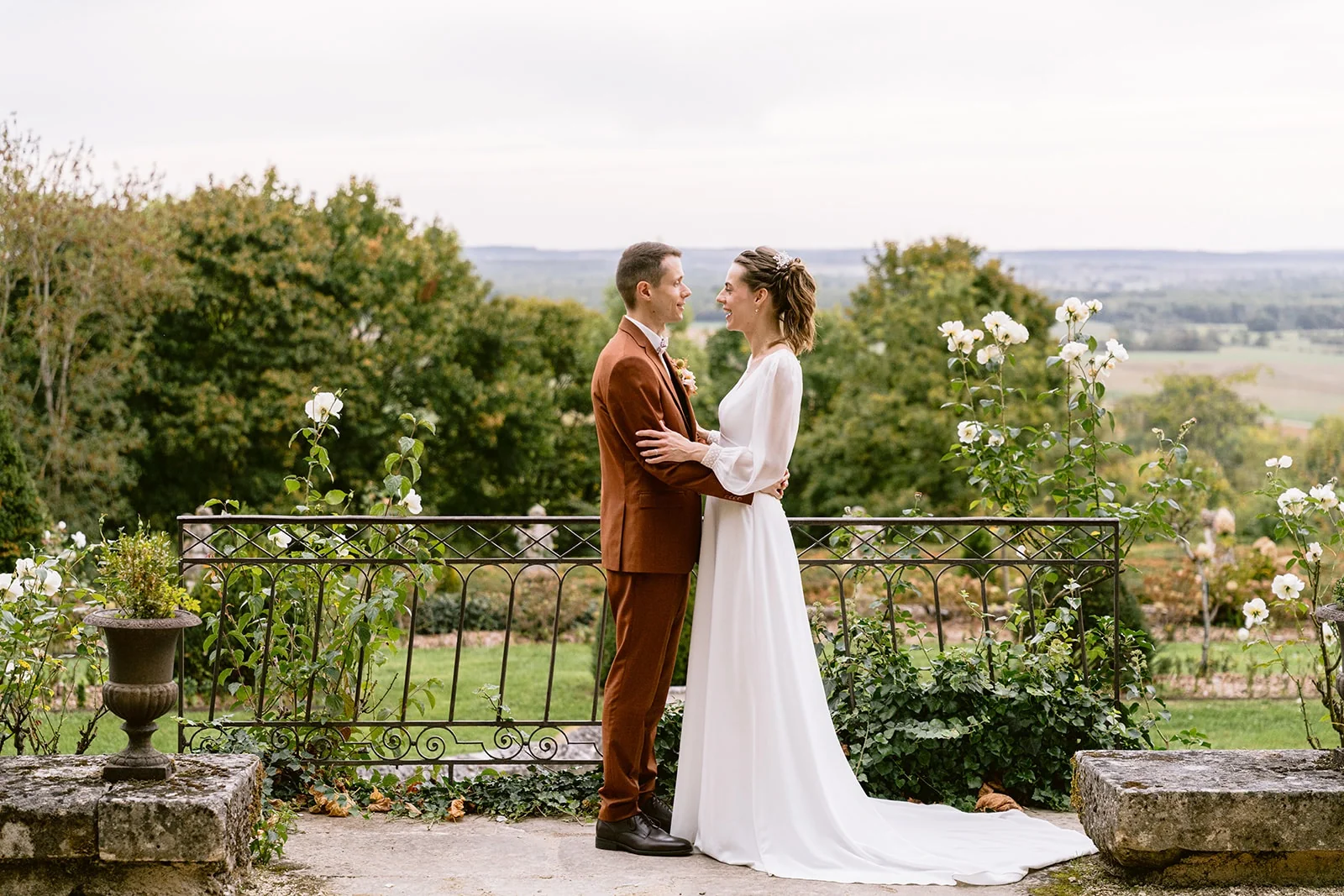 Les mariés se font face sur une terrasse bordée de rosiers, devant une balustrade et un paysage vallonné.