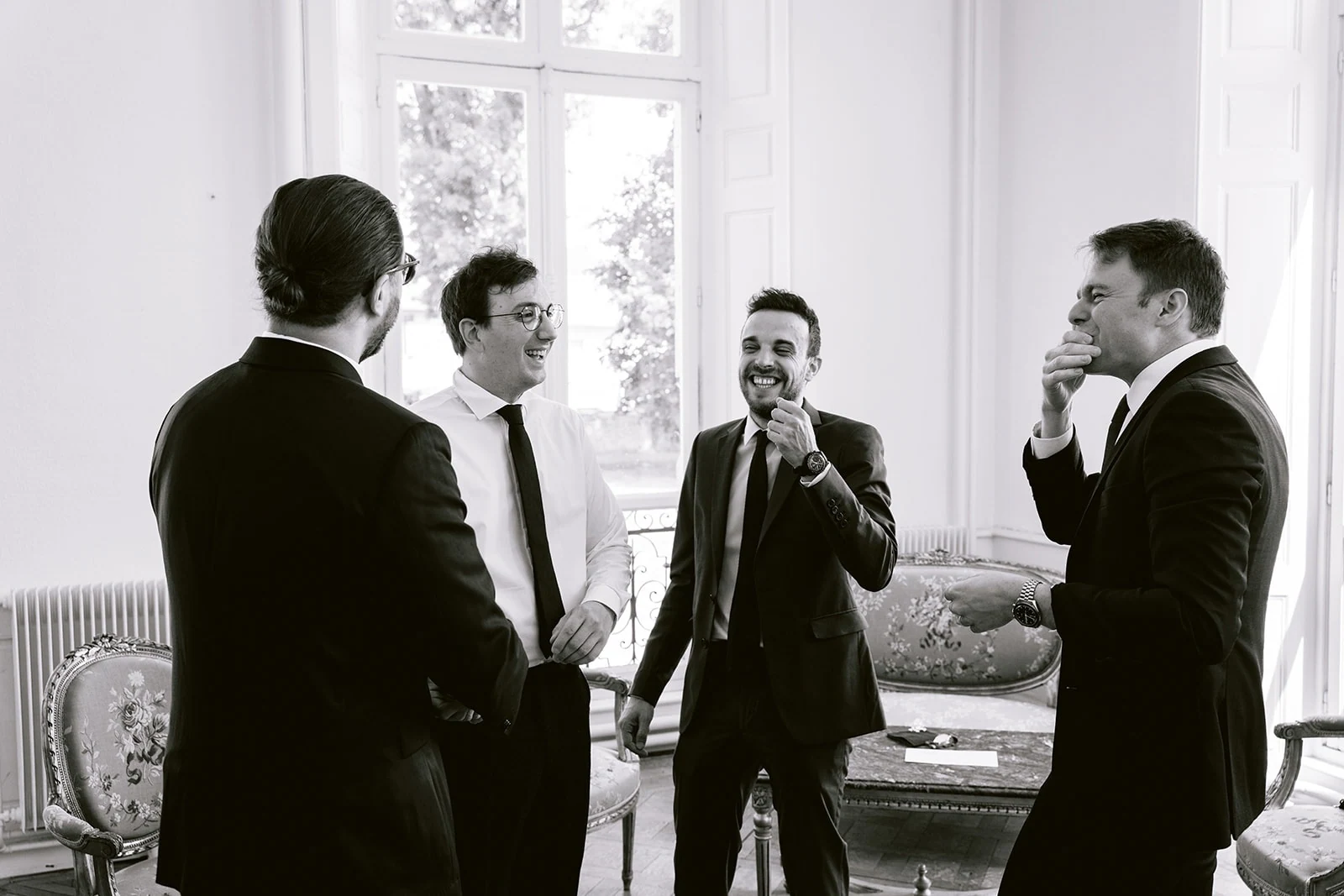 En noir et blanc, quatre hommes en tenues de cérémonie échangent des sourires dans un salon lumineux.