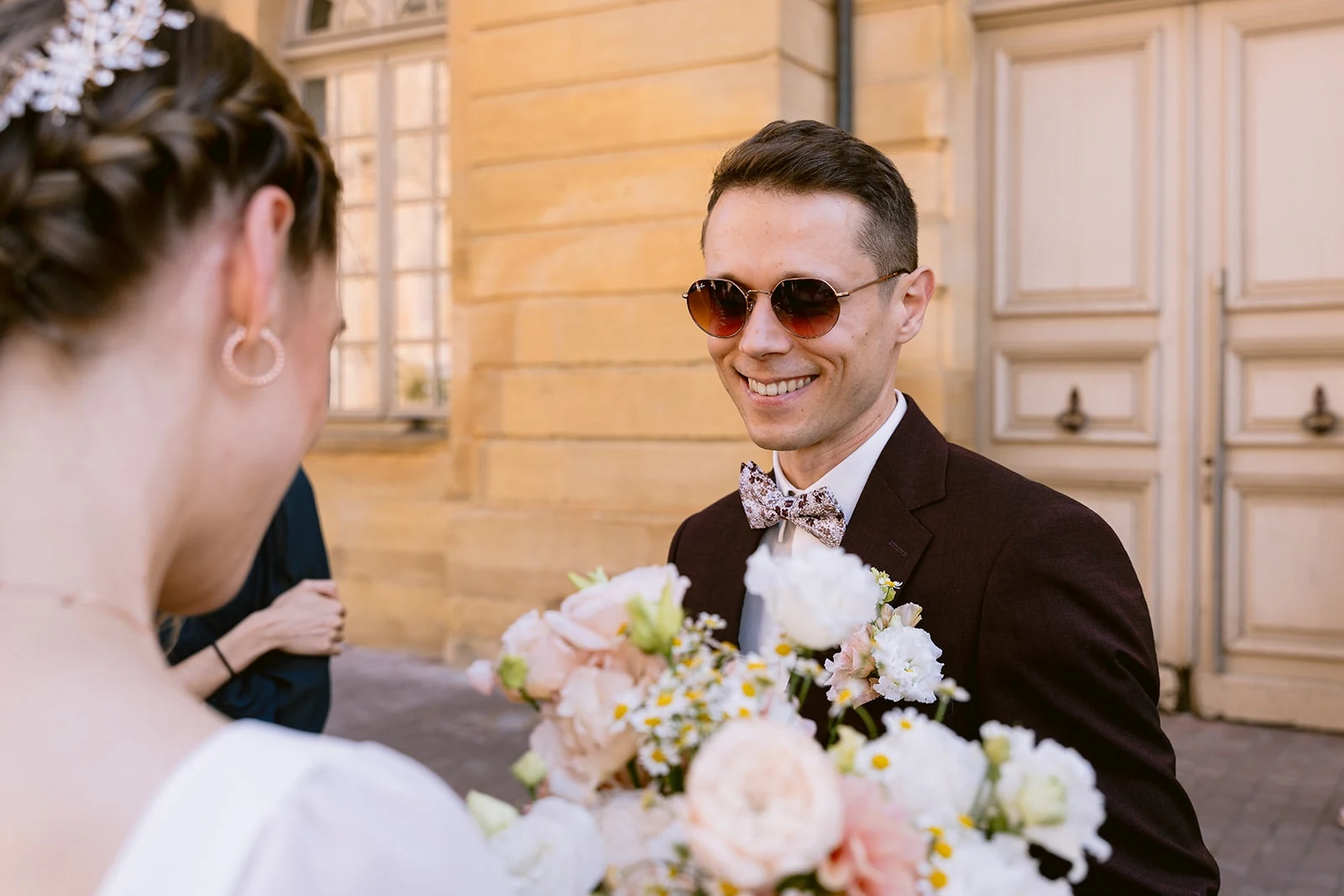 Le marié, en costume brun et lunettes de soleil, sourit face à la mariée et à son bouquet pastel.