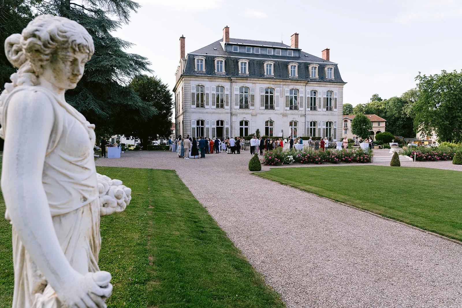 Une statue blanche borde l’allée de gravier menant à une grande demeure, où des invités sont rassemblés.