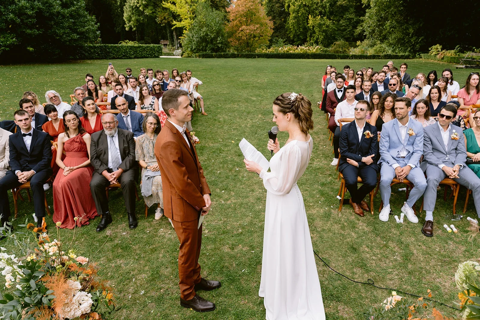 La mariée lit une feuille au micro face au marié, entourée des invités assis sur la pelouse.