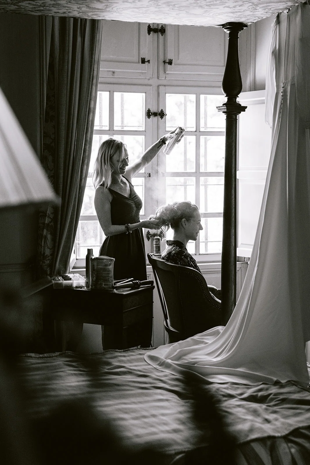 En noir et blanc, une coiffeuse vaporise de la laque sur les cheveux de la mariée, près d’une fenêtre.