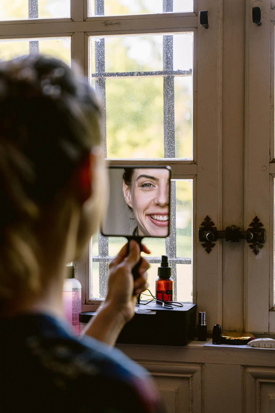 La mariée sourit à son reflet dans un petit miroir à main, près d’une fenêtre.