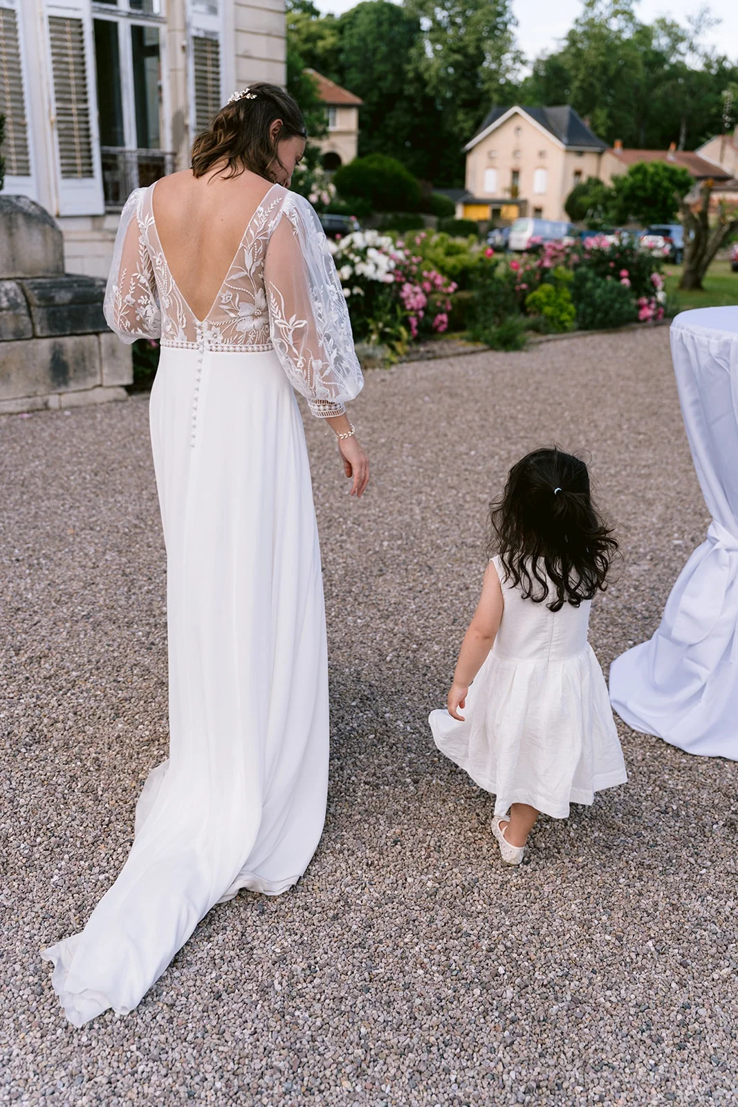 Vue de dos, la mariée marche près d’un enfant en robe blanche sur une allée de gravier bordée de fleurs.