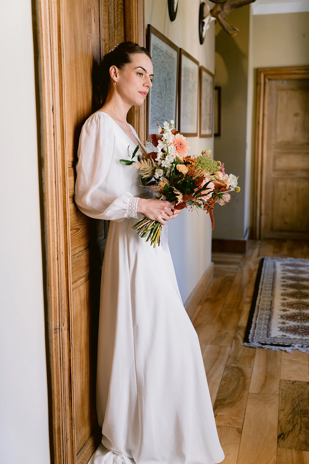 La mariée, bouquet à la main, pose de profil contre une porte en bois dans un couloir.