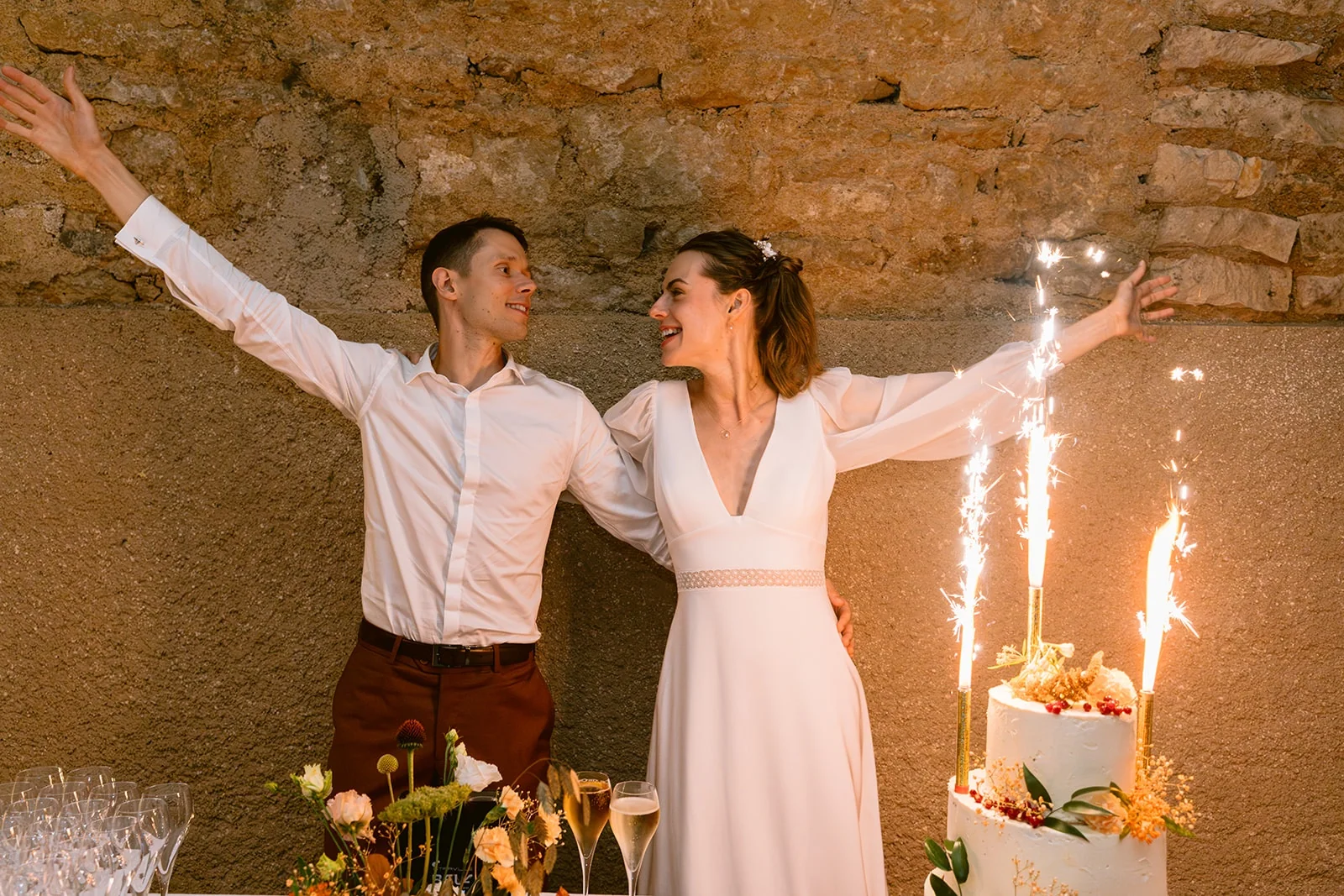 Les mariés lèvent les bras à côté de leur gâteau, illuminé par des fontaines d’étincelles.
