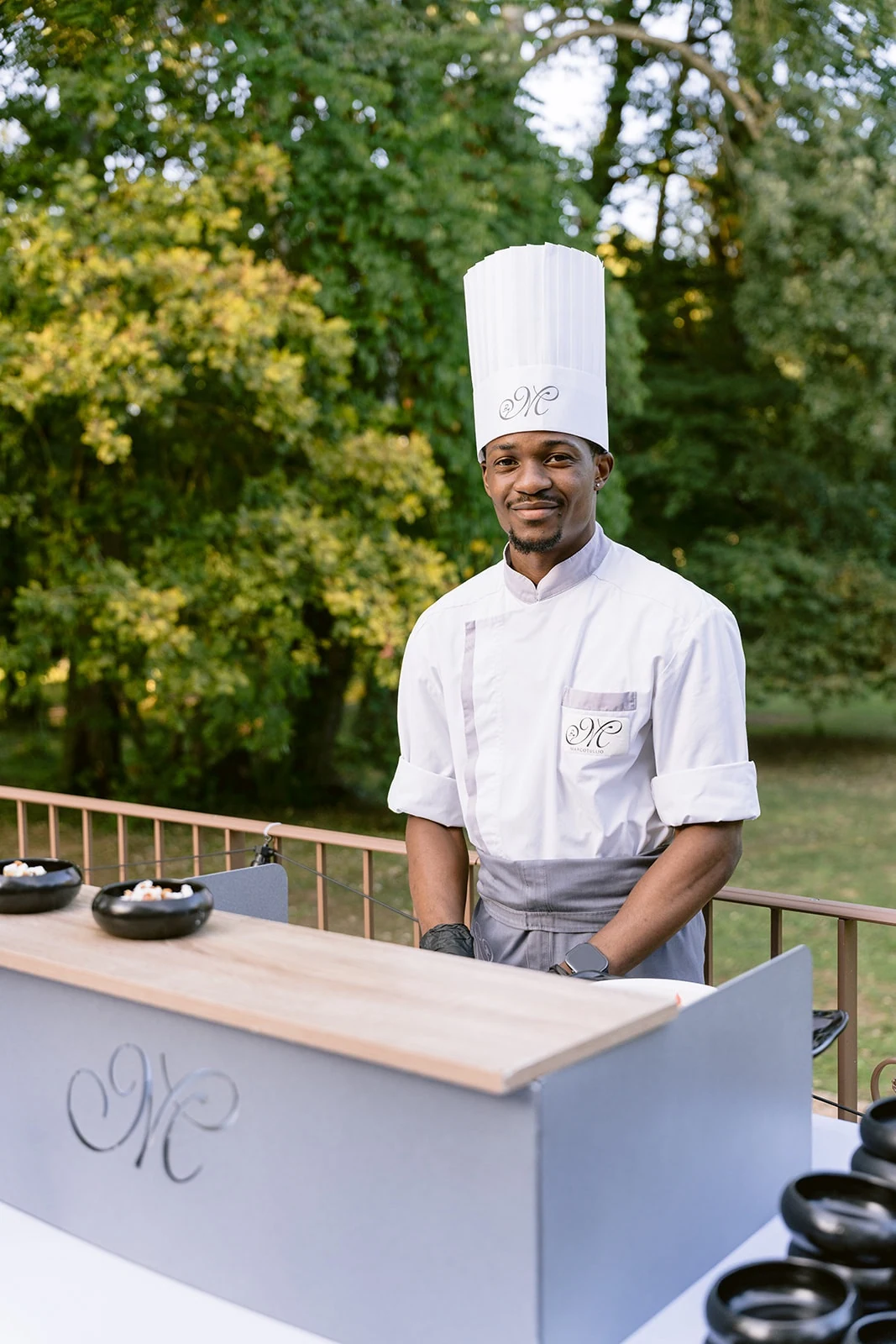 Un cuisinier en toque blanche se tient derrière son poste de préparation dans le jardin.