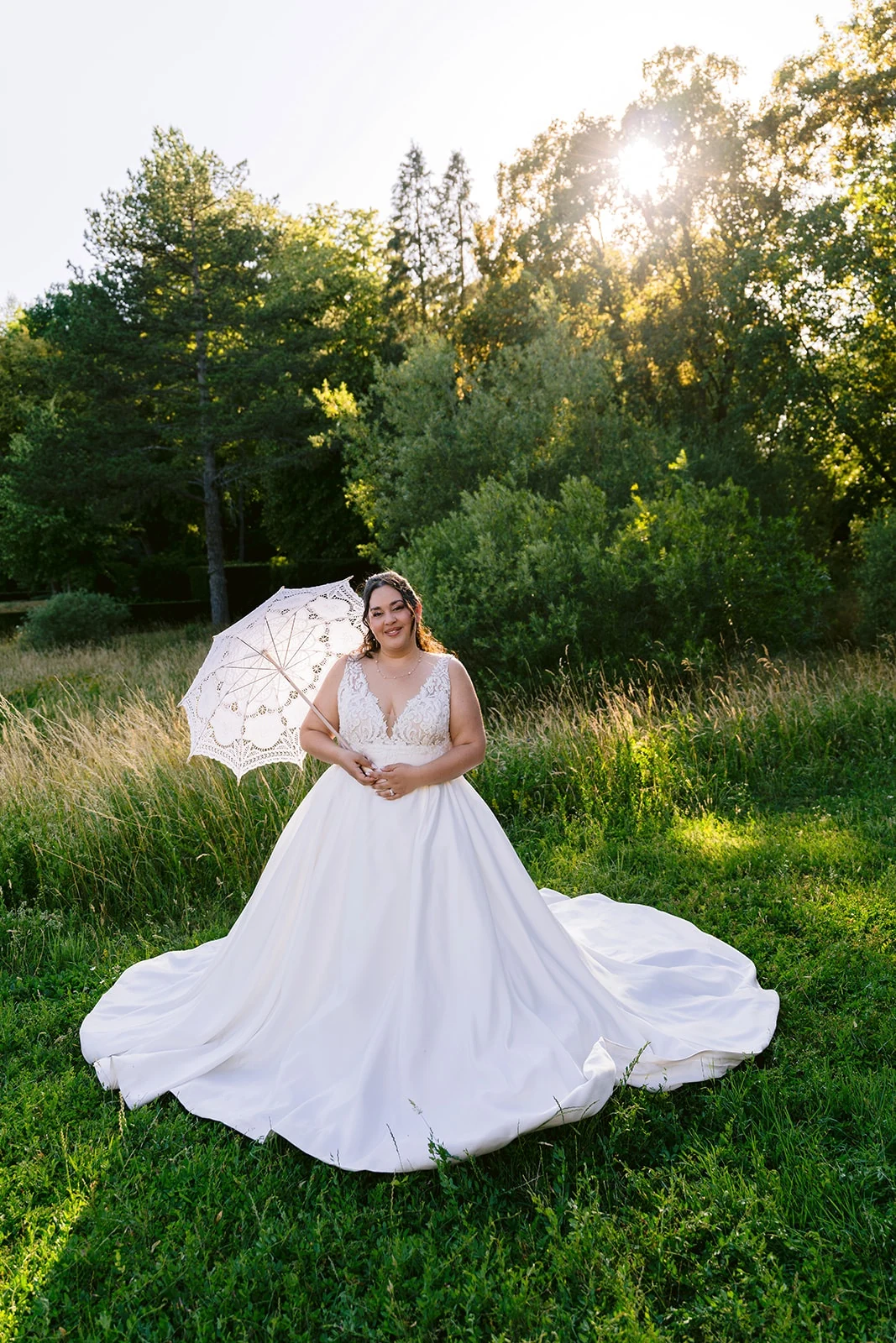 Une mariée pose avec une ombrelle blanche, sa traîne déployée dans l’herbe à la lumière du soir.