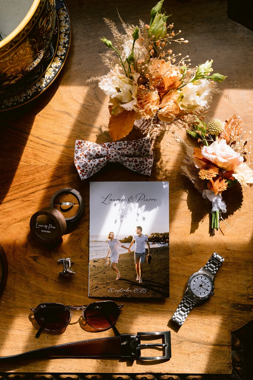 Bouquet, boutonnière, nœud papillon fleuri, montre, lunettes de soleil et ceinture sont disposés sur une table en bois.