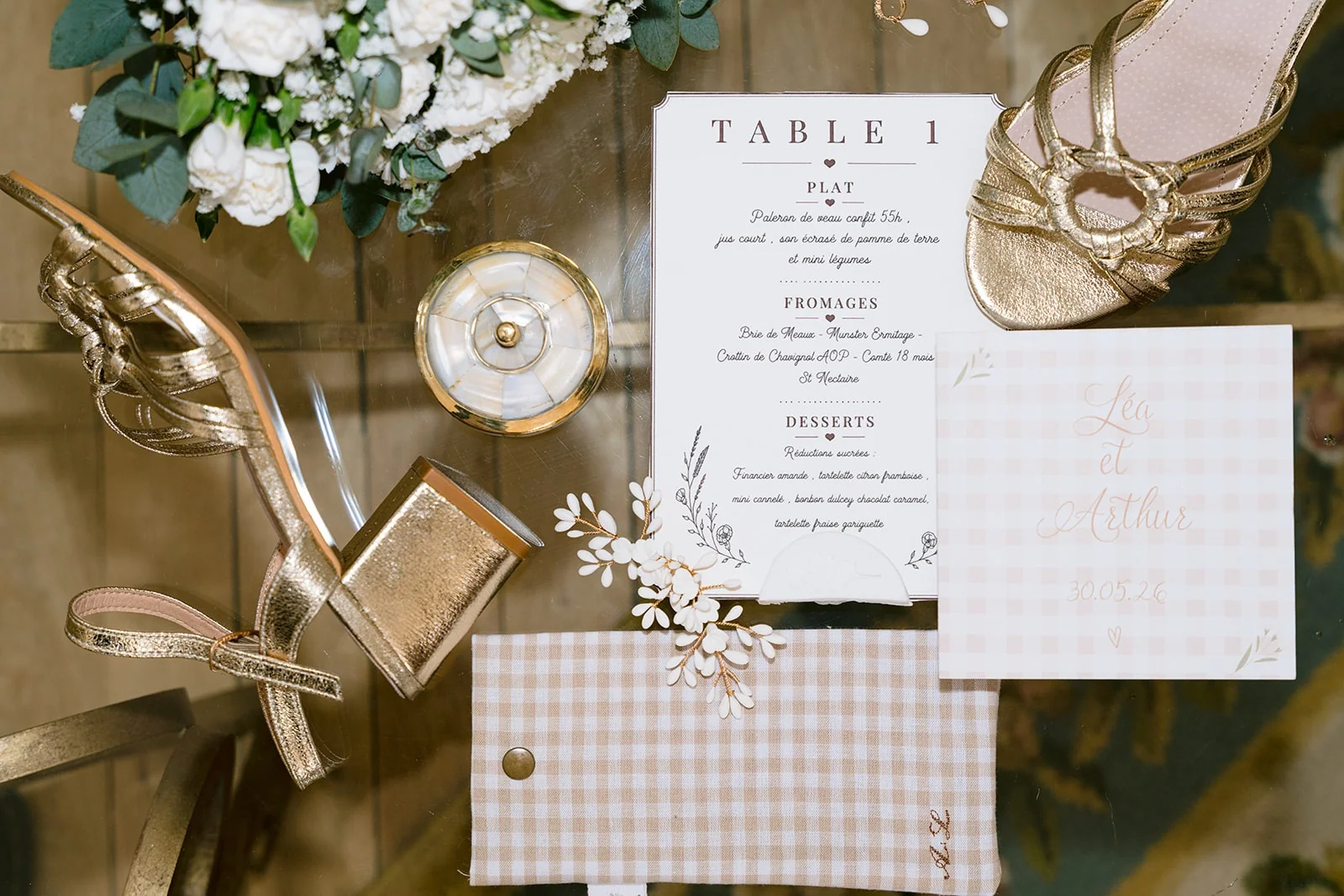 Un menu de réception, des sandales dorées et la papeterie du mariage disposés près d’un bouquet blanc.