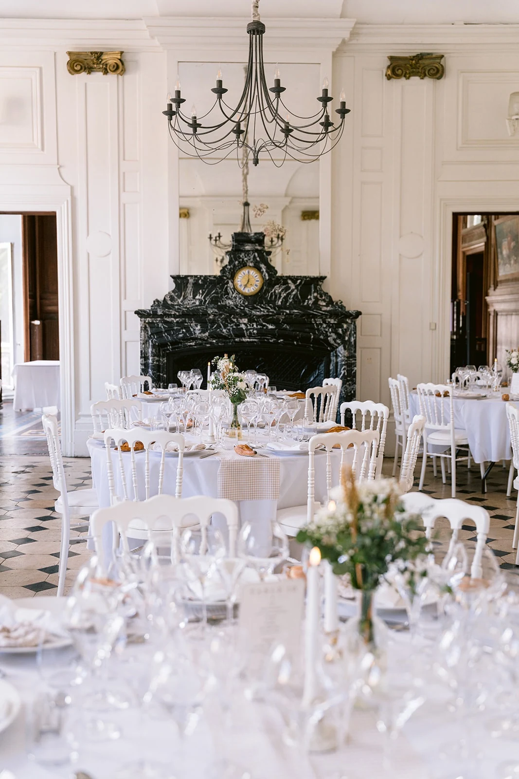 Un salon de réception aux tables blanches fleuries, dominé par un lustre et une cheminée sombre surmontée d’une pendule.