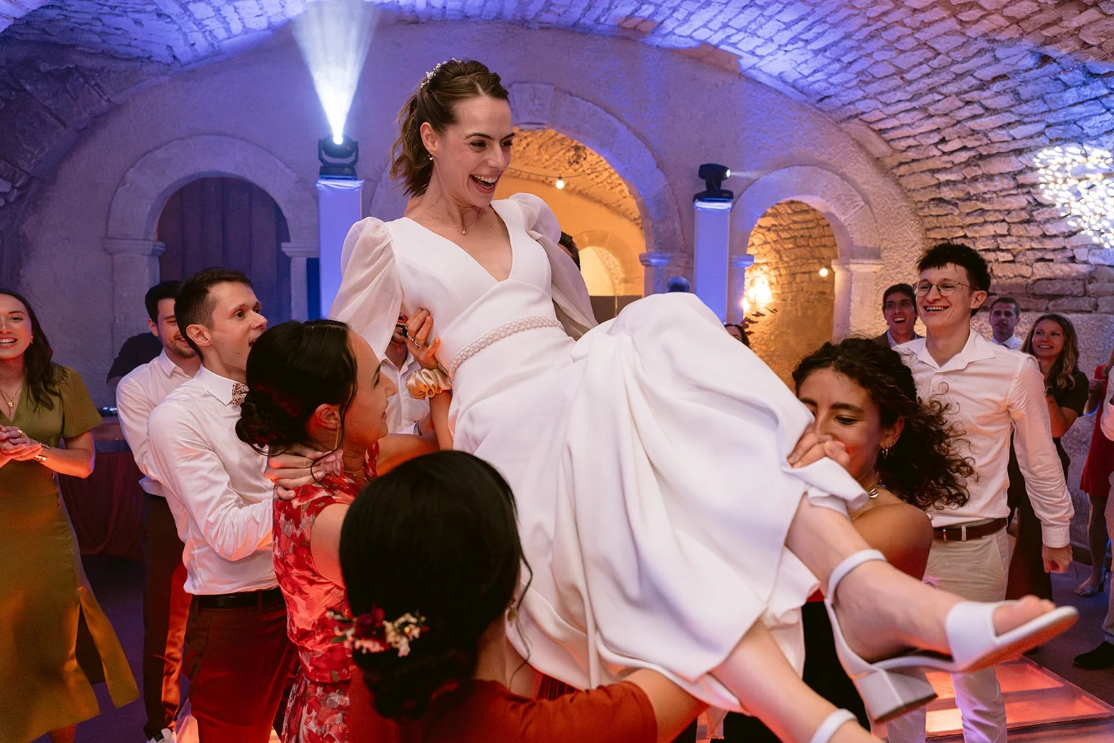 La mariée rit tandis que plusieurs invités la portent au-dessus de la piste de danse.