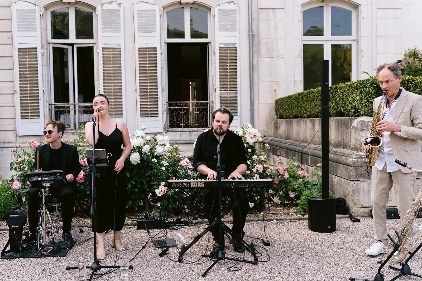 Une chanteuse et trois musiciens, dont un saxophoniste, jouent devant la façade fleurie d’une demeure.