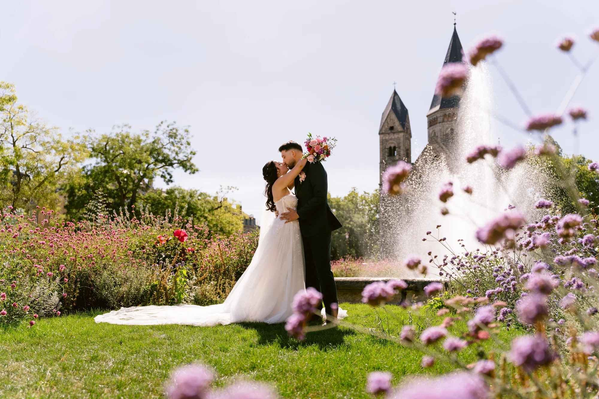 Les mariés s’embrassent dans un jardin, entre les fleurs et les tours en pierre.