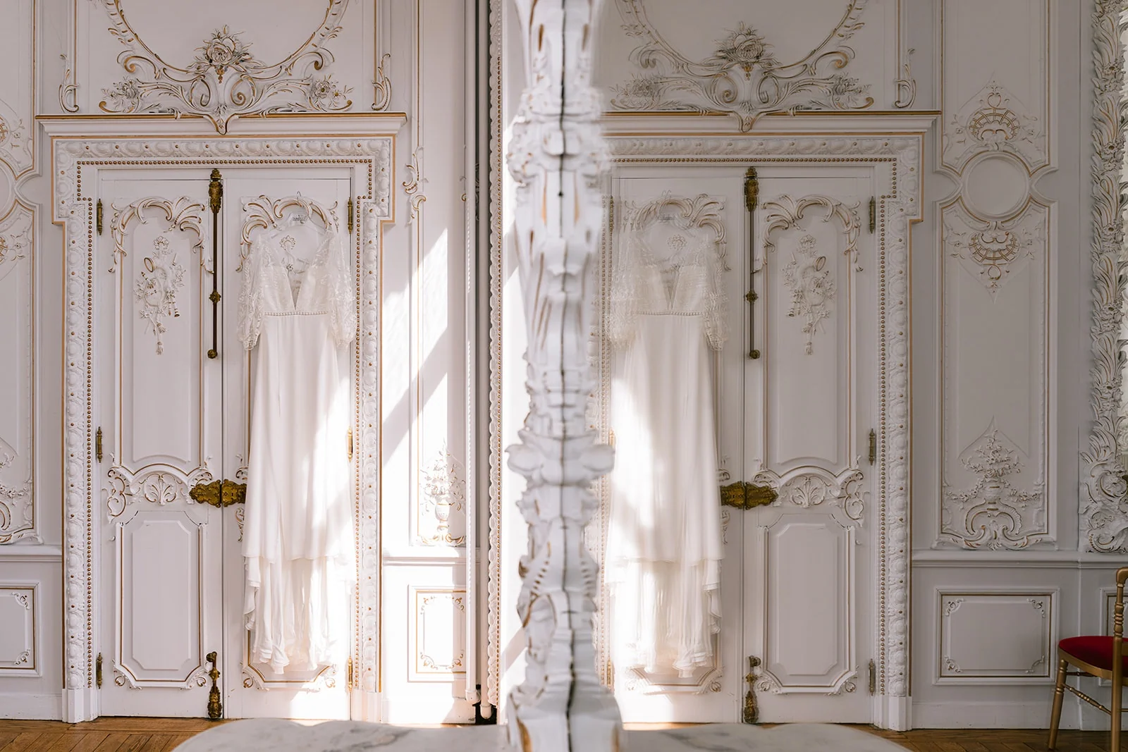 Une robe de mariée suspendue à une porte moulurée se reflète dans un grand miroir, traversée par la lumière du soleil.