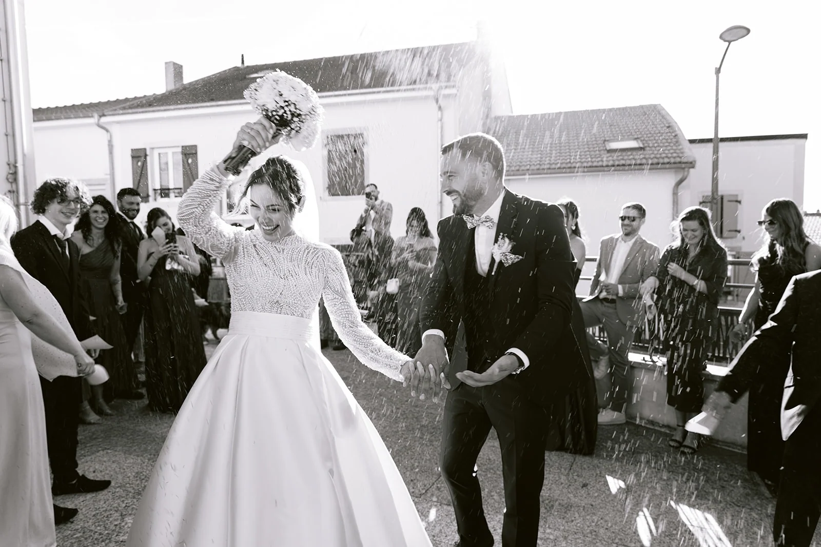 En noir et blanc, les mariés sortent de l’église sous les confettis et lèvent leurs mains jointes.