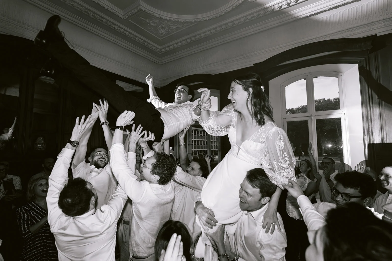 En noir et blanc, les mariés sont soulevés par leurs invités au milieu de la fête.