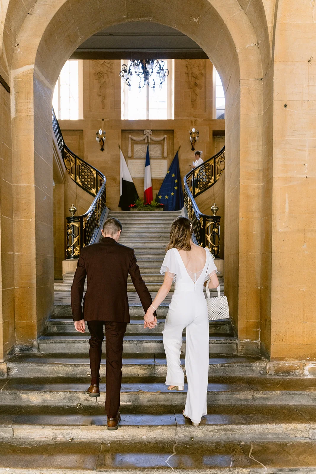 Vus de dos, les mariés montent les marches d’un bâtiment de pierre en se tenant la main.