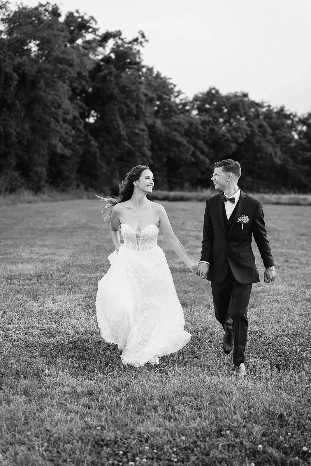En noir et blanc, les mariés marchent main dans la main dans une prairie.