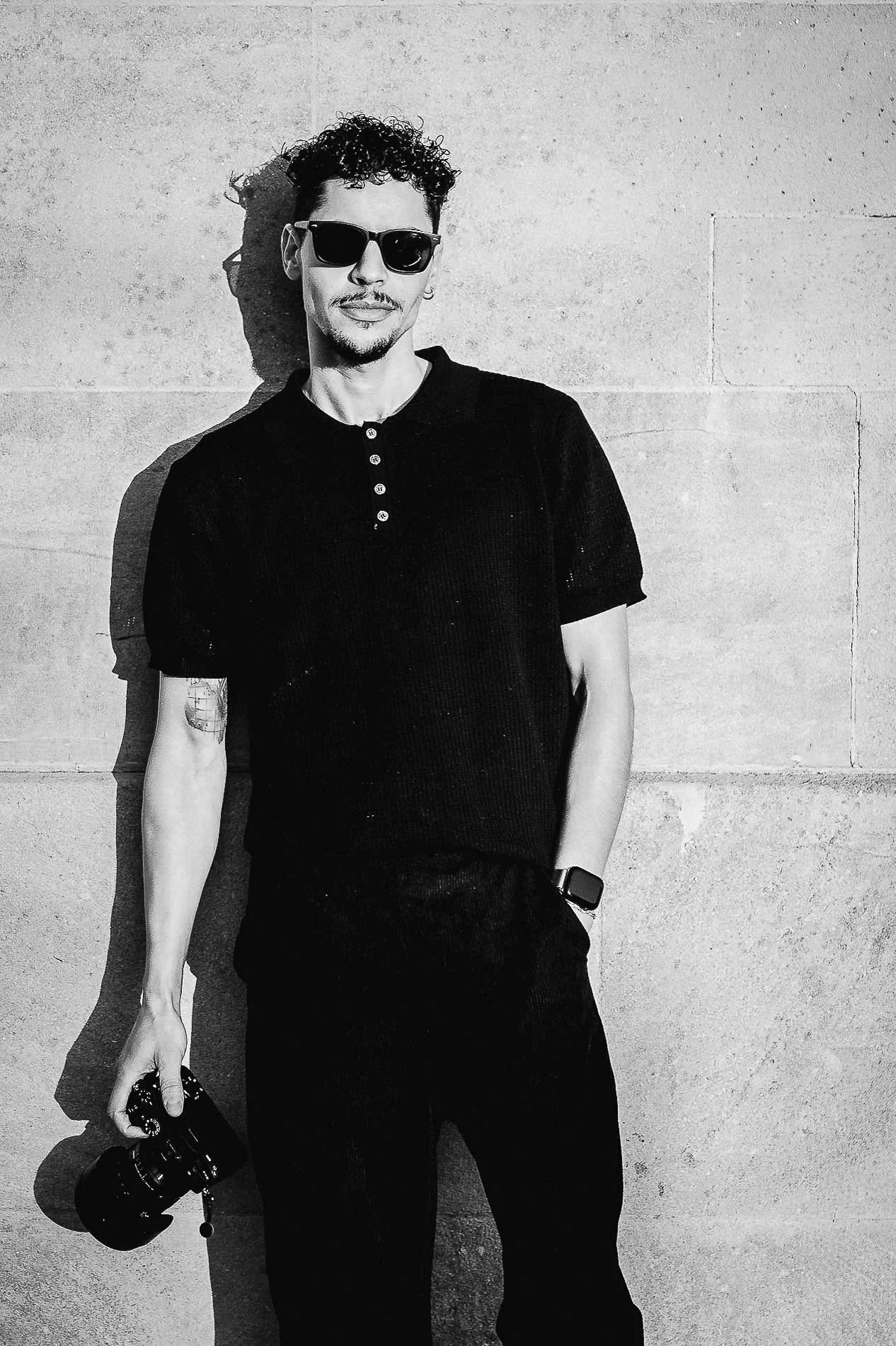 Portrait en noir et blanc du photographe, lunettes de soleil et appareil à la main, contre un mur de pierre.