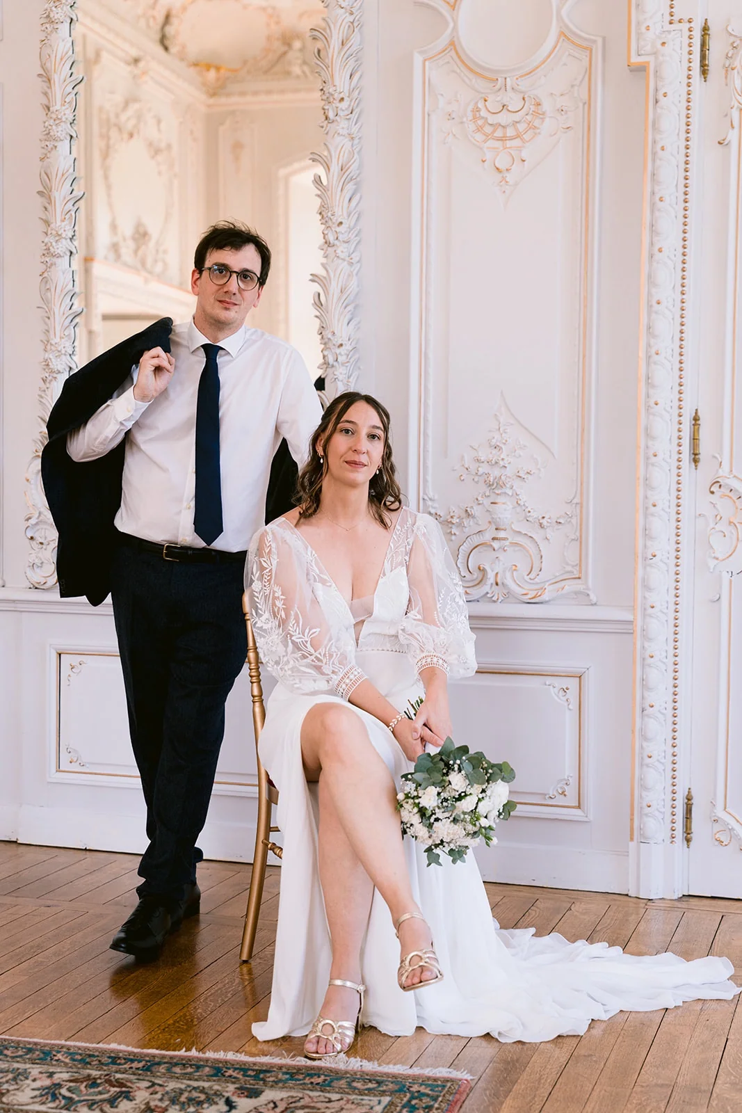 La mariée est assise avec son bouquet, à côté du marié debout, veste sur l’épaule, dans un salon aux moulures blanches.