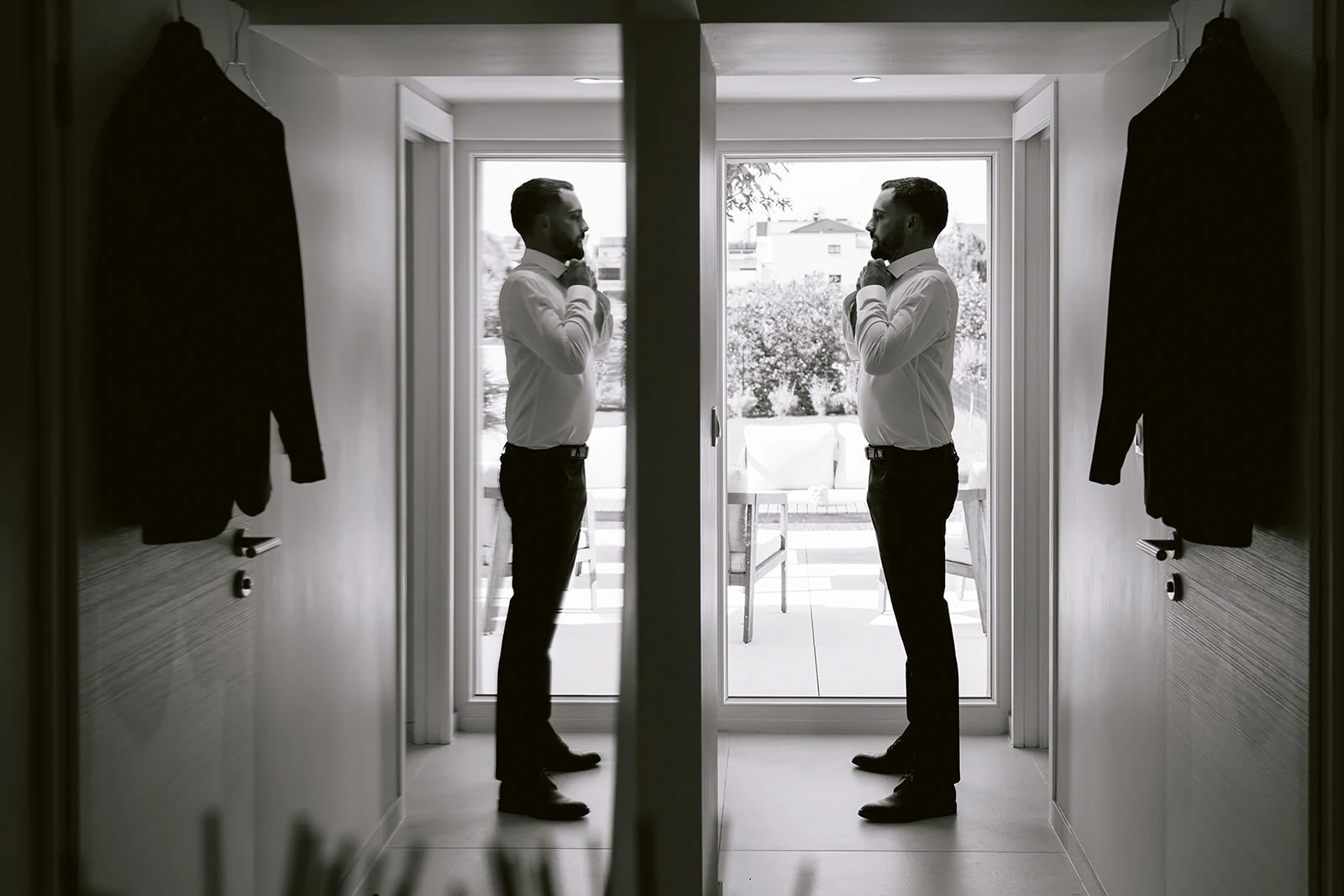 En noir et blanc, le marié ajuste sa tenue près d’une fenêtre.