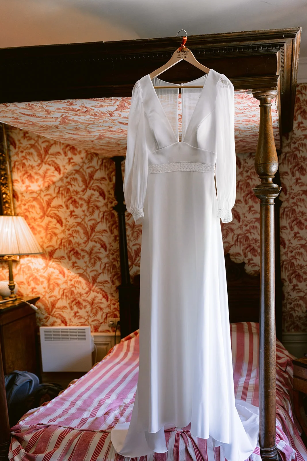 Une robe de mariée blanche à manches longues est suspendue au-dessus d’un lit à baldaquin.
