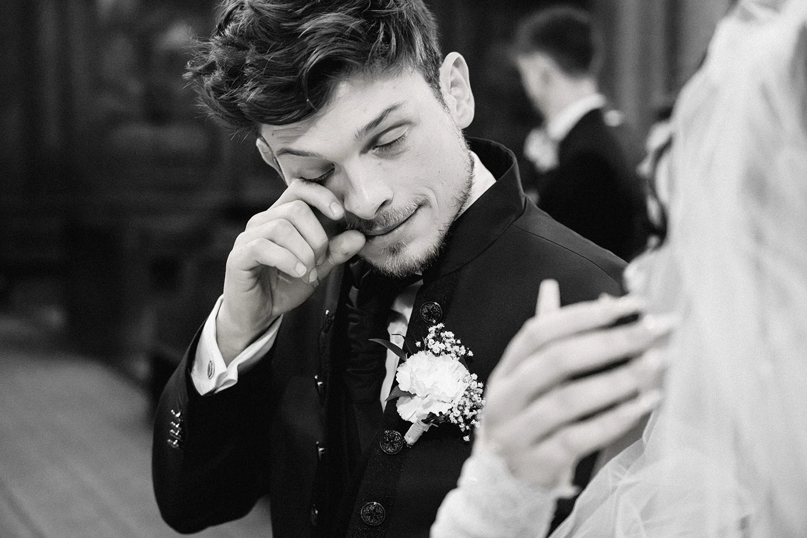 En noir et blanc, le marié essuie une larme, une main posée sur l’épaule de la mariée.