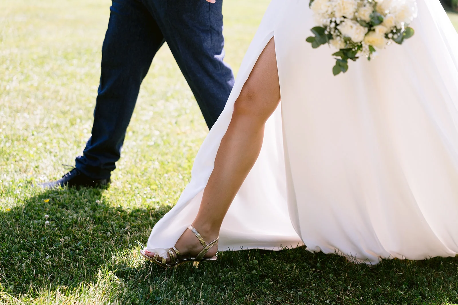 Détail des pas des mariés dans l’herbe, avec la jupe fendue de la robe, une sandale dorée et le bouquet blanc.
